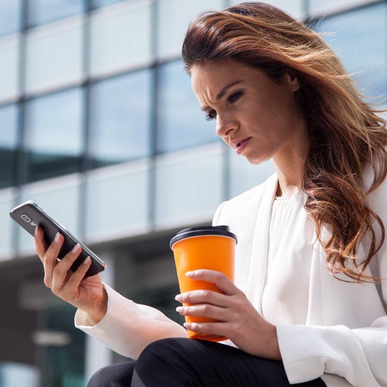 Eine junge Frau mit einem Kaffee in der Hand sitzt am Buerogebaeude auf einer Treppe und schaut irritiert auf ihr Smartphone.