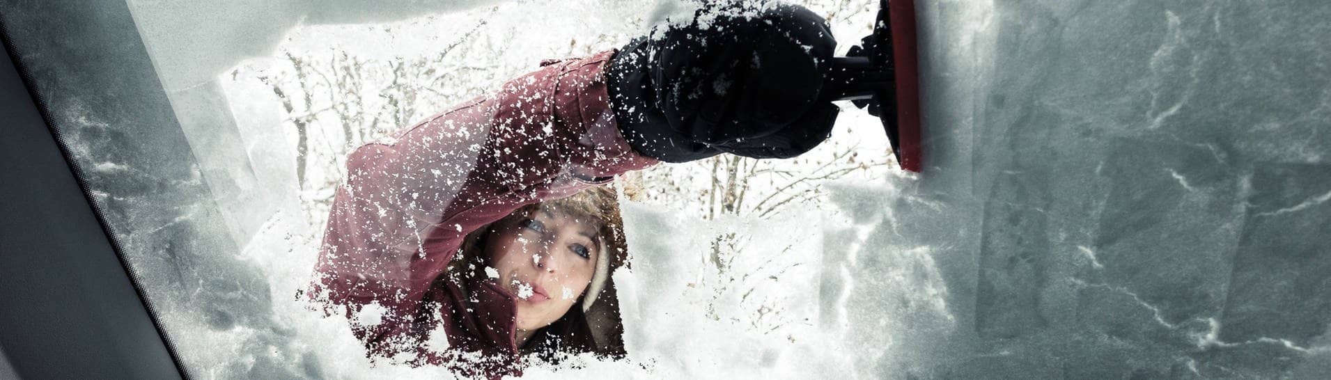 Junge Frau mit Enteiser, sie kratzt die verschneite und gefrorene Scheibe des Autos im Winter frei.