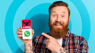 Ein Mann mit Bart hält vor blauem Hintergrund ein Handy in der Hand und zeigt darauf. Auf dem Bildschirm sind die Logos von SWR3 und Whatsapp zu sehen, da SWR3 nun auch einen Whatsapp-Kanal hat.