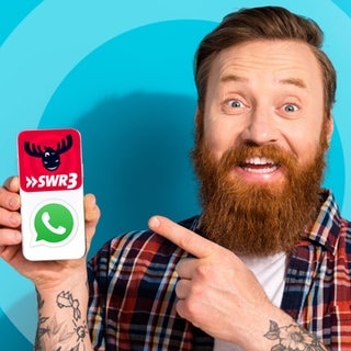 Ein Mann mit Bart hält vor blauem Hintergrund ein Handy in der Hand und zeigt darauf. Auf dem Bildschirm sind die Logos von SWR3 und Whatsapp zu sehen, da SWR3 nun auch einen Whatsapp-Kanal hat.