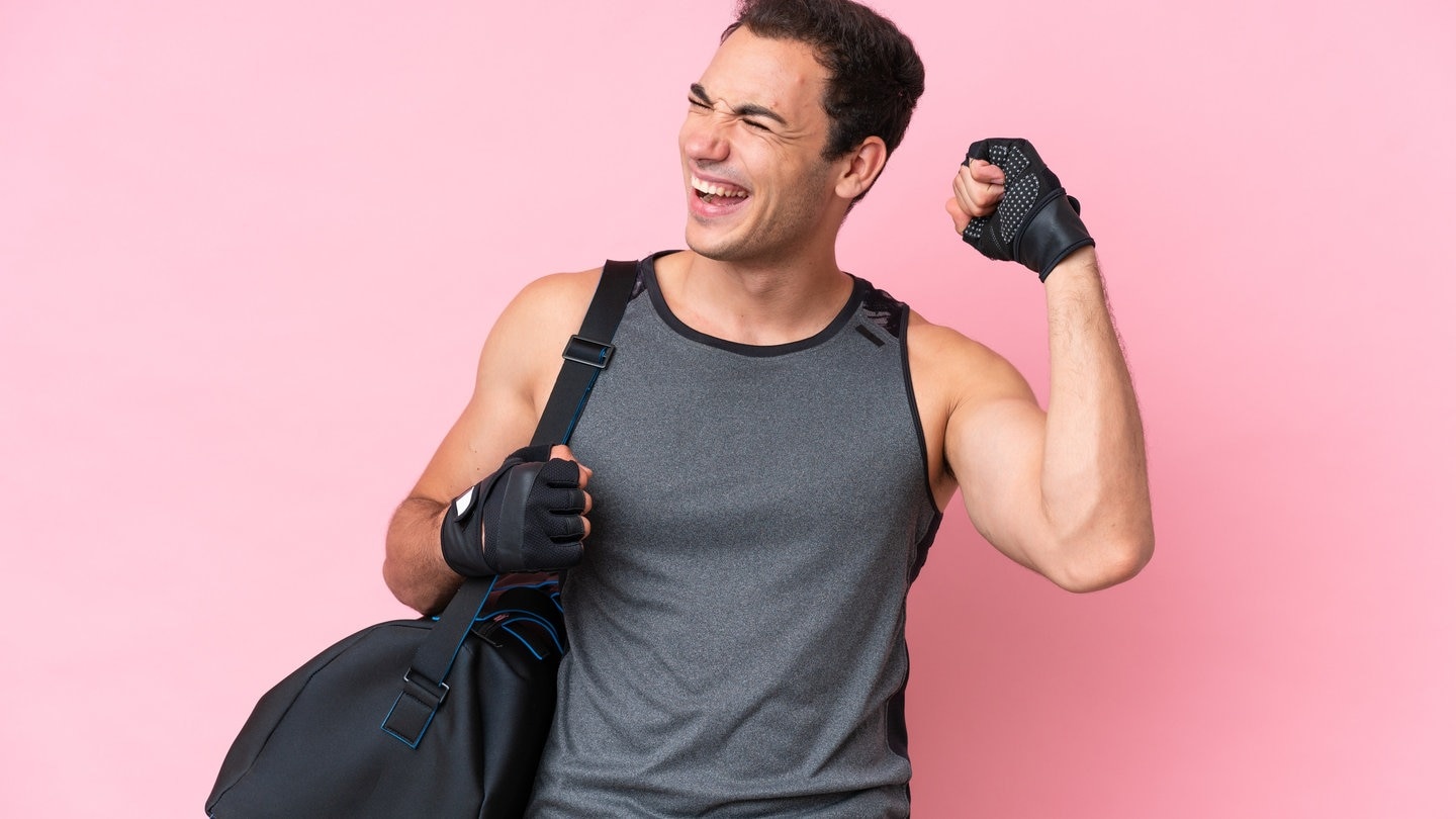 Gute Vorsätze durchhalten braucht Routine: Dieser junge Mann freut sich, dass er regelmäßig zum Sport geht, er hat eine Sporttasche bei sich und spannt den Oberarmmuskel an