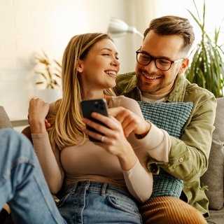 Junges Paar sitzt auf der Couch, liest auf dem Smartphone, lacht und freut sich über gute Nachrichten.