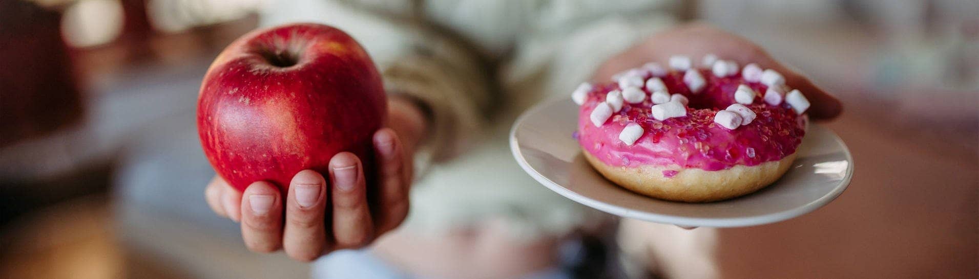 Frau hält einen Apfel und eine Donut. Beide enthalten Zucker und werden den Glukosespiegel ansteigen lassen