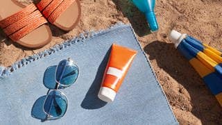 Sonnecremetube liegt auf Strandtuch mit Sonnenbrille und Sonnenschirm