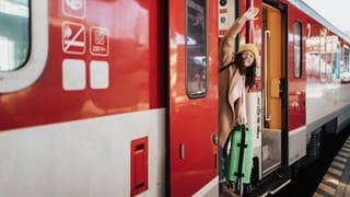 Das 49-Euro-Ticket startet jetzt. Symbolbild: Eine Frau winkt an der Tür eines Zug stehend mit grünem Koffer in der Hand