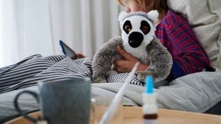 Ein Fieberthermometer, Nasenspray und eine Tasse Tee stehen vor einem Bett, in dem ein krankes Kind mit einem Kuscheltier sitzt und auf ein Tablet schaut. 