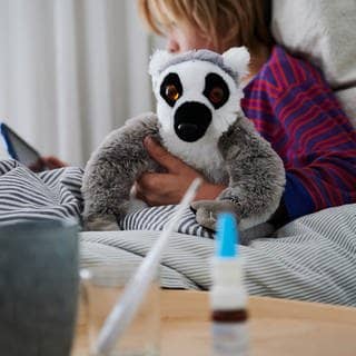 Ein Fieberthermometer, Nasenspray und eine Tasse Tee stehen vor einem Bett, in dem ein krankes Kind mit einem Kuscheltier sitzt und auf ein Tablet schaut. 
