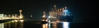 Zwei Schiffe liegen vor einem beleuchteten Hafen. Das Frachtschiff „Polesie“ wird in der Nacht von zwei Schleppern an den Kai der Seebäderbrücke in Cuxhaven gezogen.
