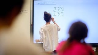 Eine Lehrerin schreibt eine Mathematikaufgabe auf eine digitale Schultafel im Klassenraum einer 4. Klasse einer Grundschule.