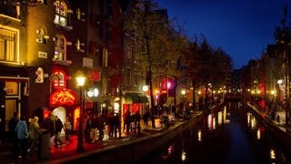 Menschen schlendern in Amsterdam am Abend an einer Gracht entlang durch den Rotlichtbezirk De Wallen.