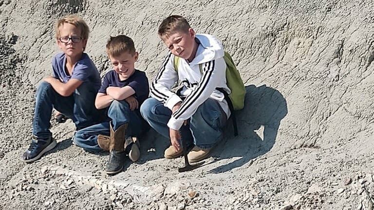 Die drei Kinder Kaiden Madsen, Liam Fisher und Jessin Fisher sitzen neben dem T-Rex-Beinknochen, den sie gefunden haben
