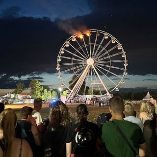 Man sieht Besucher des Highfield-Festivals, die auf ein Riesenrad blicken, an dem Flammen zu sehen sind. Auf dem Highfield-Festival am Störmthaler See ist ein Feuer an einem Riesenrad ausgebrochen.