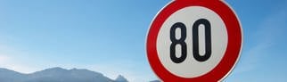 Ein Tempo-80-Schild steht an einer Straße. Im Hintergrund ist ein Motorradfahrer.