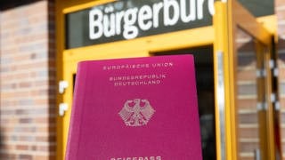 Eine Person hält einen Reisepass vor einem Bürgerbüro in der Hand.