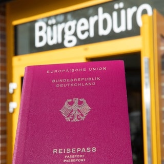 Eine Person hält einen Reisepass vor einem Bürgerbüro in der Hand.