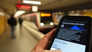 Ein Fahrgast hält ein Smartphone mit einem digitalen Deutschlandticket an einer U-Bahnstation in der Hand.