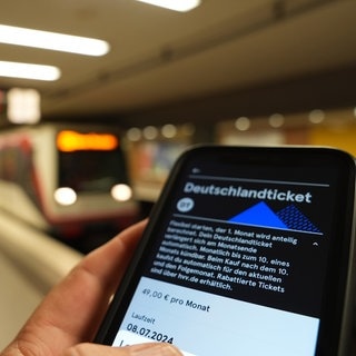 Ein Fahrgast hält ein Smartphone mit einem digitalen Deutschlandticket an einer U-Bahnstation in der Hand.