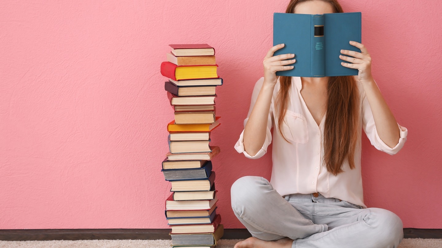 Eine Frau sitzt auf dem Boden und liest ein Buch, neben ihr ist ein großer Bücherstapel.