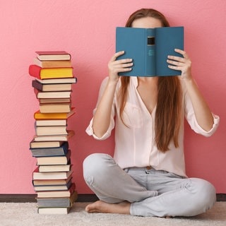 Eine Frau sitzt auf dem Boden und liest ein Buch, neben ihr ist ein großer Bücherstapel.