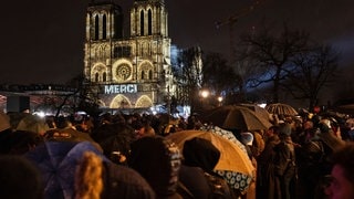 Zuschauer versammeln sich draußen vor der Kathedrale Notre Dame in Paris. Auf die Kathedrale werden Lichter und das Wort „merci“ projiziert.