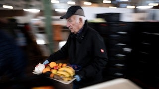 Lange Schlangen vor der Tafel: Ein älterer Mitarbeiter trägt eine Tablet mit Obst.