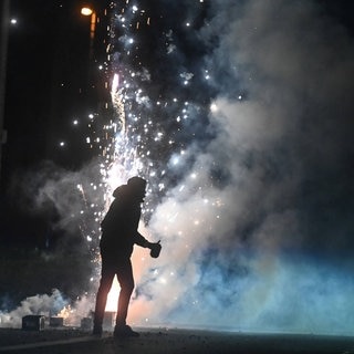 Ein Mann steht an Silvester vor Feuerwerk mit einem Böller in der Hand