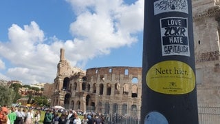 Ein „Nett hier“-Aufkleber klebt an einem Mast vorm Kolosseum in Rom.