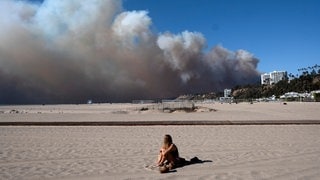 Eine Frau sitzt in Santa Monica am Strand und beobachtet, wie eine große Rauchwolke von einem Waldbrand über Pacific Palisades aufsteigt.