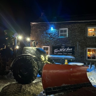 Ein Traktor mit Schneepflug steht vor dem Pub Tan Hill Inn im Schnee