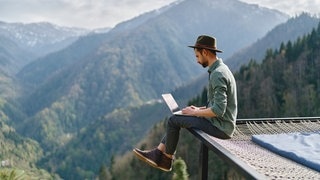 Ein Mann sitzt mit seinem Laptop auf dem Schoß vor einem Bergpanorama