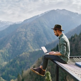 Ein Mann sitzt mit seinem Laptop auf dem Schoß vor einem Bergpanorama