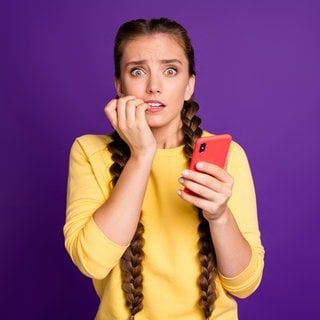 Eine junge Frau beißt auf den Fingernägeln und hält ihr Handy. (Symbolbild für Angst vorm Telefonieren)