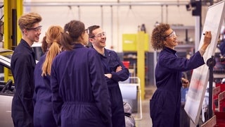 Symbolbild Auszubildende, man sieht mehrere junge Leute in einer Fabrihalle lachend vor einer Tafel stehen, auf der eine junge Frau etwas einträgt.