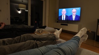 Drei Menschen liegen auf einem Sofa, während auf dem Fernseher Olaf Scholz (SPD) und Friedrich Merz (CDU) beim TV-Duell zur Bundestagswahl zu sehen sind.