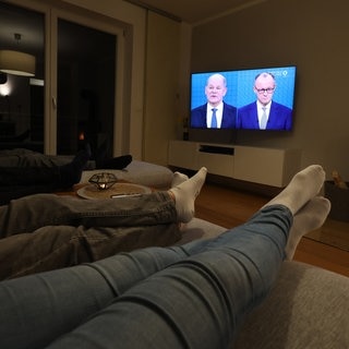 Drei Menschen liegen auf einem Sofa, während auf dem Fernseher Olaf Scholz (SPD) und Friedrich Merz (CDU) beim TV-Duell zur Bundestagswahl zu sehen sind.