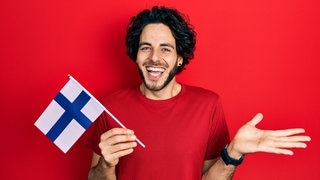 Ein junger Mann lächelt und hält eine finnische Flagge. Finnland ist wieder auf Platz 1 des World Happiness Reports.