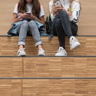 Zwei Schülerinnen sitzen mit ihrem Handy im Foyer einer Schule.