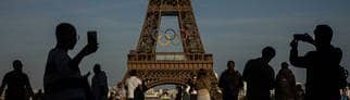 Die olympischen Ringe sind auf dem Eiffelturm in Paris zu sehen. Im Vordergrund Silhouetten von Menschen, die Selfies machen.