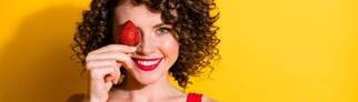 Junge Frau mit braunen Locken hält sich eine rote Erdbeere vors Gesicht und lacht. Symbolbild für Wissen und Fakten rund um Erdbeeren.