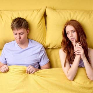 Junges Paar liegt im Bett. Sie schaut skeptisch zu ihm rüber, er ist verunsichert, weil er keine Lust hat auf Sex.