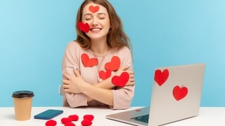 Junge Frau, beklebt mit Herzen, sitzt verliebt an einem Schreibtisch mit Laptop.