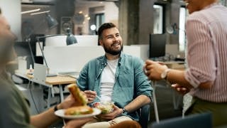 Drei Leute, die wohl miteinander arbeiten, machen gemeinsam eine Pause, so wie es im Gesetz verordnet ist. Sie essen etwas im Büro und lachen miteinander.