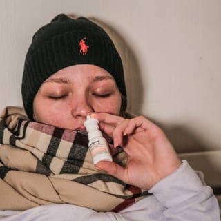 Kranke Frau mit Schal und schwarzer Mütze nutzt ein Nasenspray