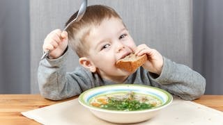 Kleiner Junge löffelt Hühnersuppe und steckt sich ein Stück Brot in den Mund. Das hilft sicher gegen Erkältung.