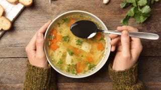 Hühnersuppe oder Hühnerbrühe kann bei Erkältung helfen. Hier im Bild ein Teller mit Suppe, auf dem Tisch liegen noch Kräuter und Brot, man sieht Frauenhände. In einer Hand hält sie einen Löffel.