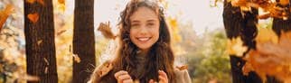 Herbstbild einer jungen Frau, die im Wald und im Sonnenschein in die Kamera lächelt. Um sie herum fallen Herbstblätter auf den Boden. Symbolbild für die vielen Gründe, warum der November eingentlich ein ganz toller Monat ist. 
