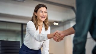 Junge Frau im Business-Outfit, schüttelt die Hand von ihrem Gegenüber. Sie freut sich über Tipps zur Beförderung und einen Aufstieg im Job.