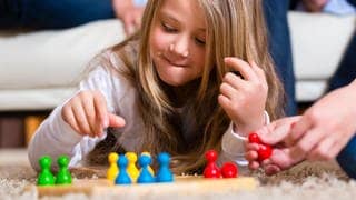 Kleines Mädchen spielt „Mensch ärgere dich nicht“, vor ihr ist das Spielfeld und in der Hand hält sie eine Spielfigur.
