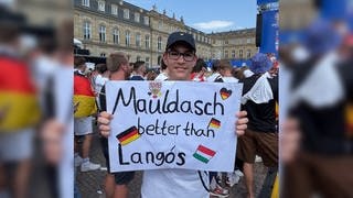 Ein junger männlicher Fußballfan in Deutschland-Trikot hält ein Schild hoch. Auf dem Schild steht: „Mauldasch better than Langós“