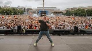 SWR3-Reporter Josh Kochhann steht auf der Bühne vor der Menschenmenge beim Public Viewing auf dem Schlossplatz in Stuttgart.